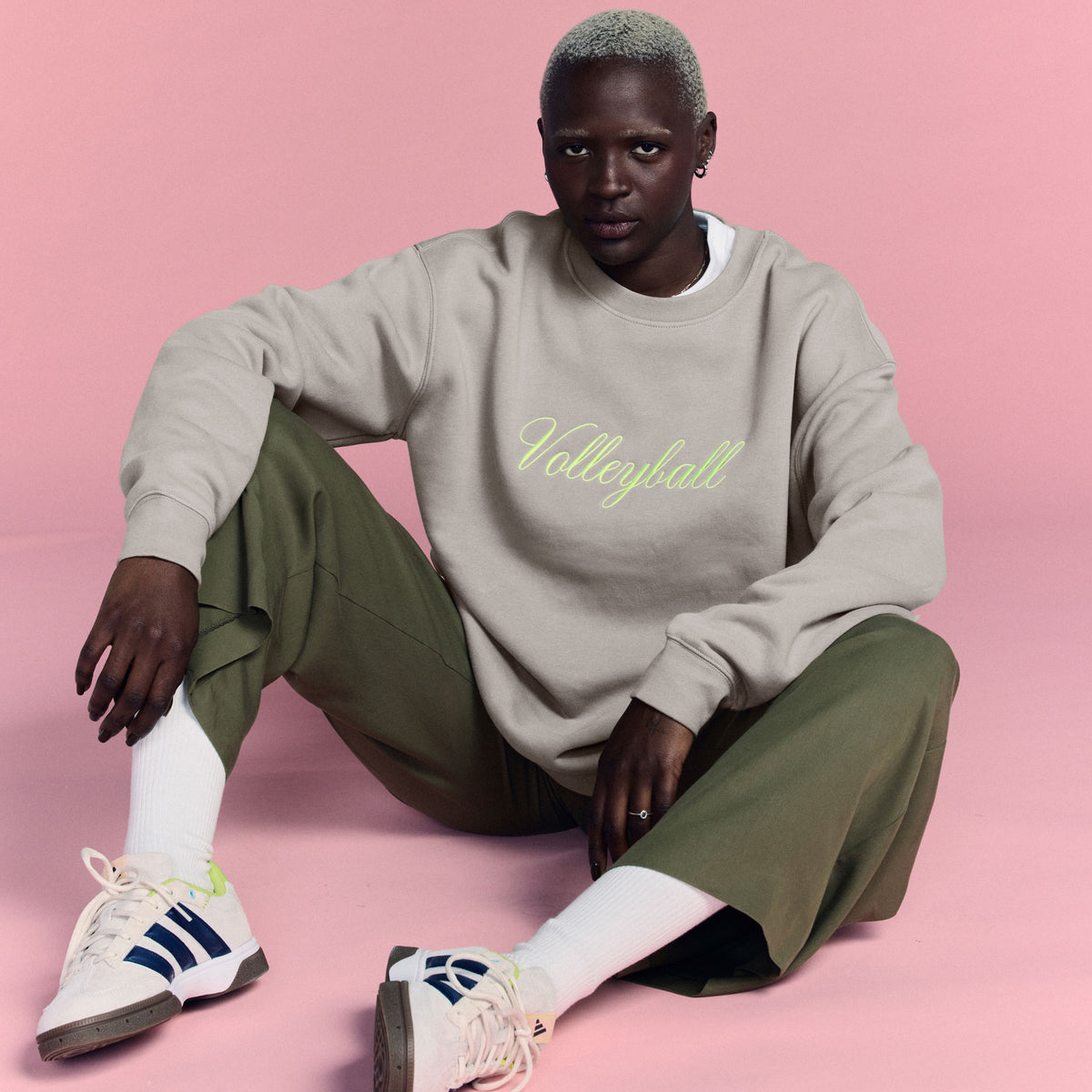 Person wearing a beige sweatshirt with 'Volleyball' text, green pants, and white sneakers on a pink background